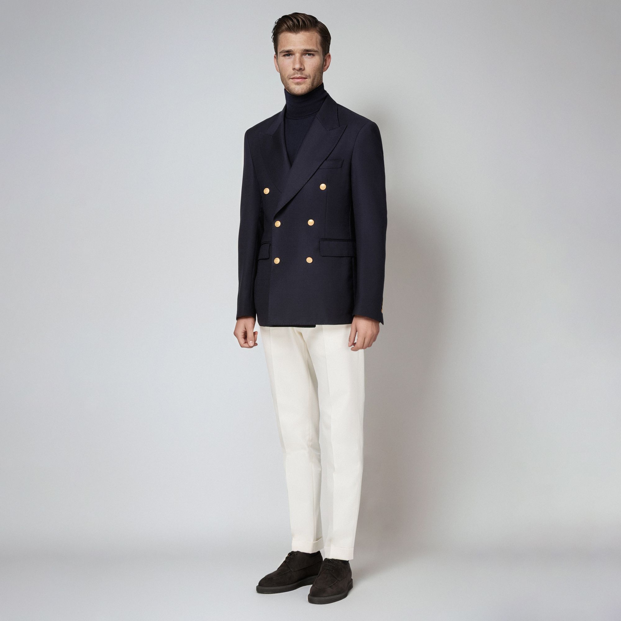 Man in an Elio Evan total look with a navy double-breasted blazer with gold buttons, navy turtleneck and white trousers.